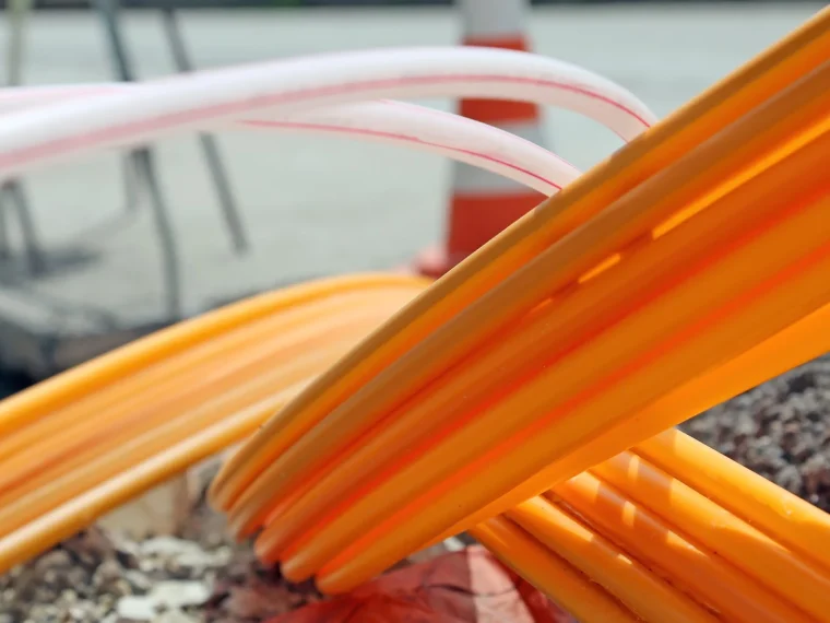 Close-up van oranje en witte kabelbuizen boven een open sleuf, met een verkeerskegel op de achtergrond