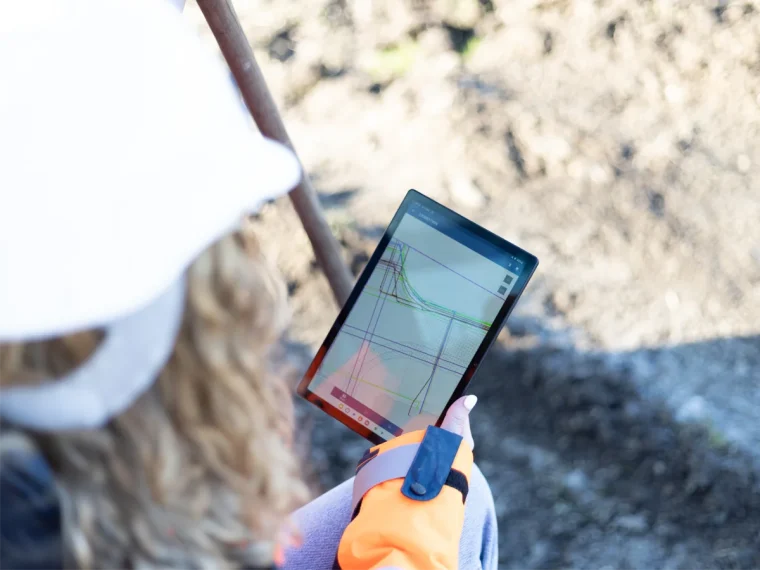 Vrouw met witte helm bekijkt een kaart met kabels en leidingen op een tablet bij een graaflocatie