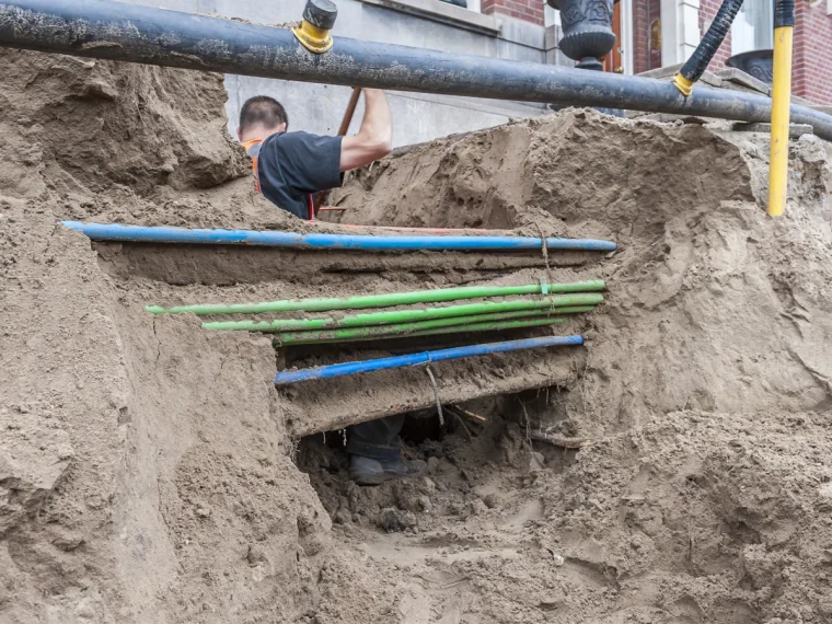 Grondwerker in een open sleuf met blauwe en groene kabels en leidingen in verschillende lagen, bij een bakstenen pand