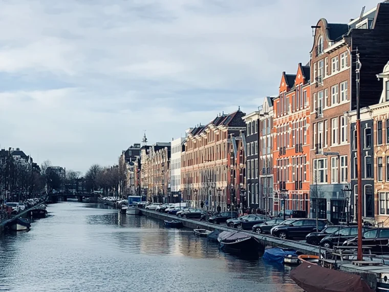 Gracht met bootjes, geparkeerde auto’s en een rij grachtenpanden onder een bewolkte lucht