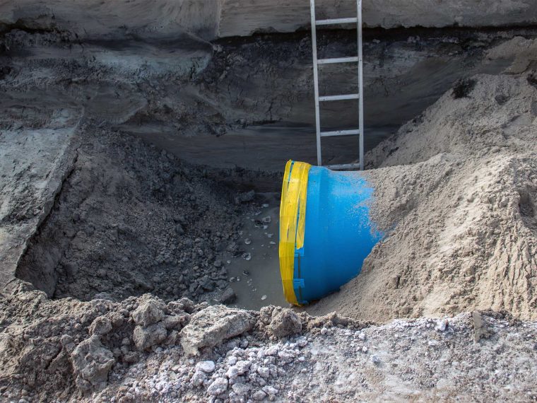 Bouwput met een blauwe leiding met gele tape, natte grond en een ladder tegen de wand