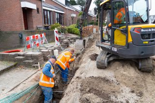 Twee grondwerkers in oranje hesjes graven in een sleuf met kabels in een woonstraat, naast een minigraver