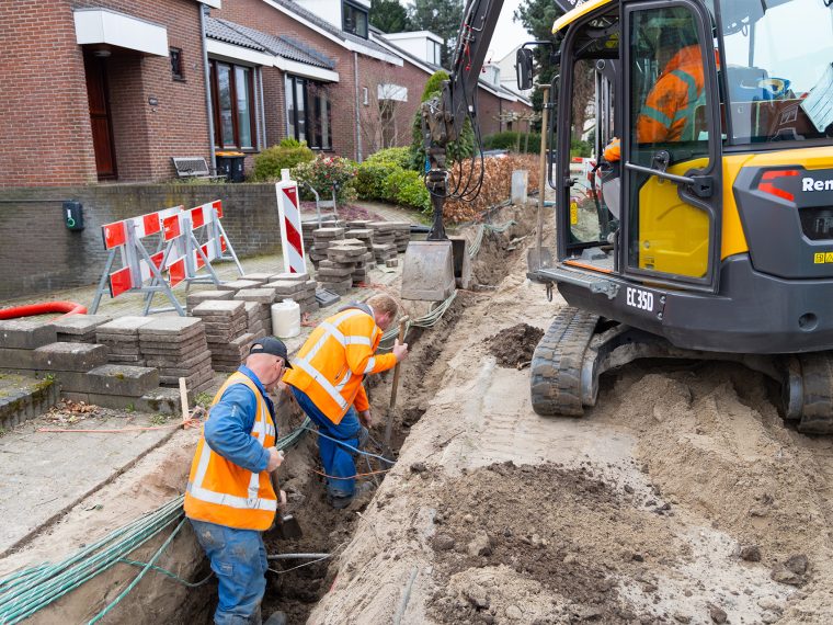 Twee grondwerkers in oranje hesjes graven in een sleuf met kabels in een woonstraat, naast een minigraver