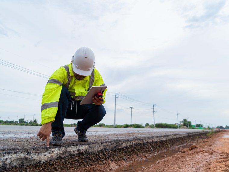 Man met helm en geel hesje zit gehurkt langs een weg en wijst naar het asfalt, met een tablet in zijn hand