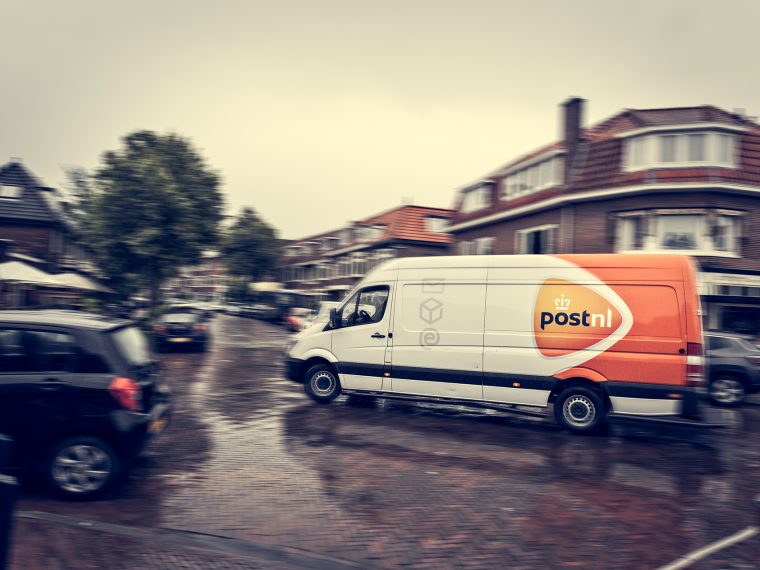 Bestelbus van PostNL rijdt door een natte woonstraat met geparkeerde auto’s
