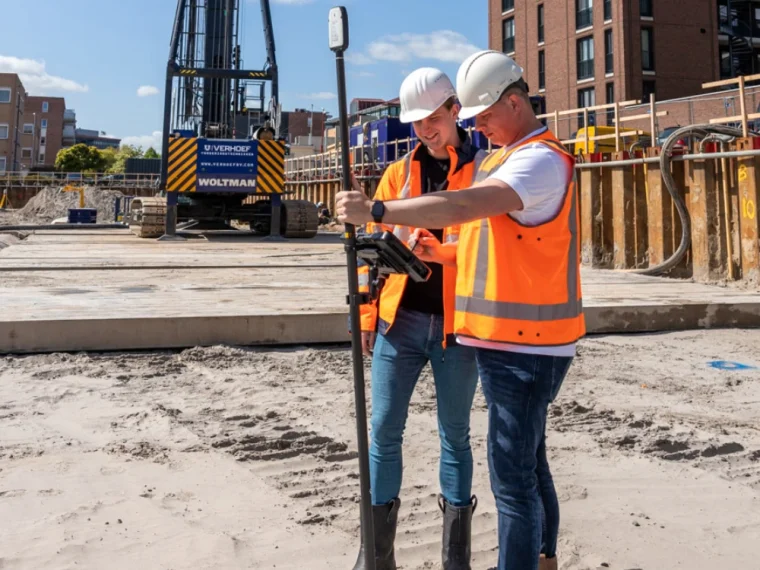 Twee medewerkers met helm en veiligheidshesje kijken op een bouwplaats samen naar een tablet op een meetstok