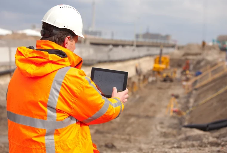 Man met helm en oranje werkjas bekijkt een tekening op een tablet bij een graaflocatie