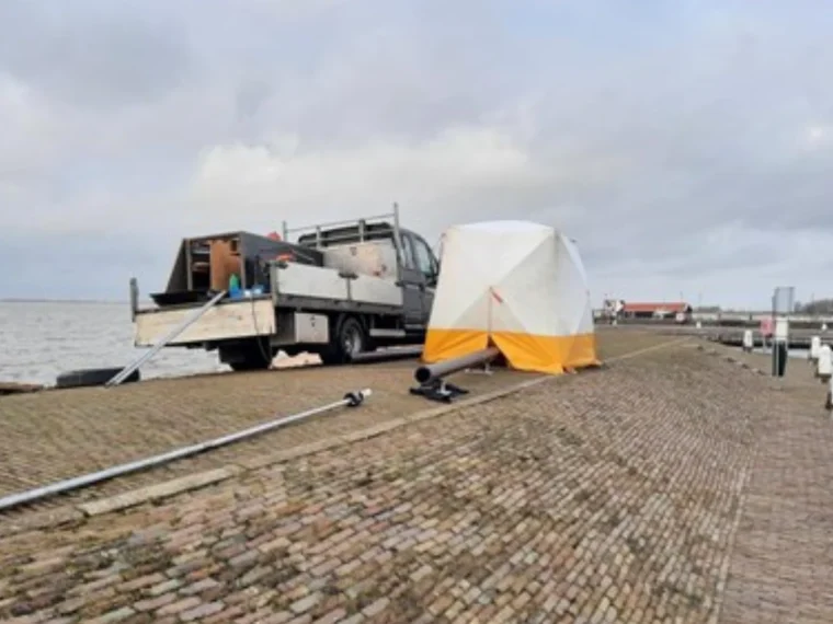 Werkbus en wit-gele werktent op een klinkerkade langs het water onder een grijze lucht
