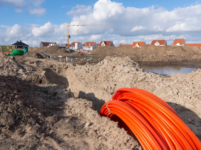 Rol oranje glasvezelbuis in een open sleuf in een nieuwbouwwijk met een bouwkraan op de achtergrond