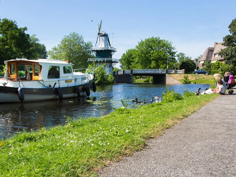 Motorboot op een vaart met een molen en een brug, naast een grasoever met een wandelpad