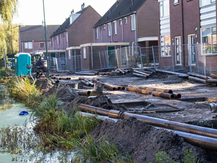 Werkzaamheden in een woonstraat met buizen op de grond, bouwhekken en een sloot op de voorgrond