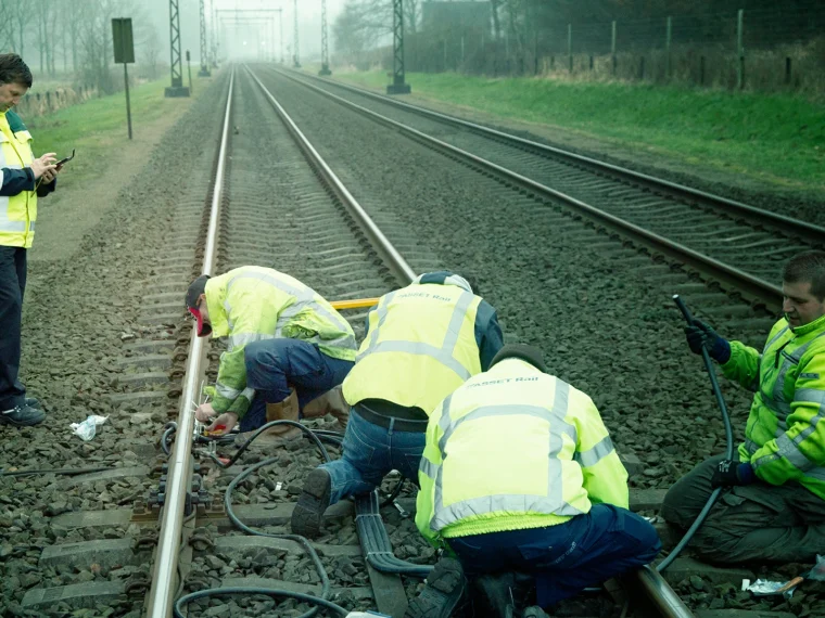 Spoorwerkers in geel veiligheidshesje werken aan het spoor, terwijl een collega op zijn telefoon kijkt