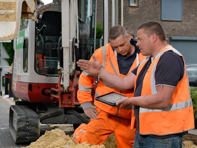 Twee grondwerkers in oranje werkkleding bekijken een tablet bij een minigraver in een woonstraat