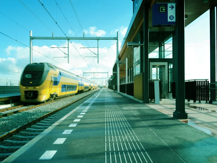 Gele trein rijdt langs een leeg perron van een modern treinstation onder een blauwe lucht