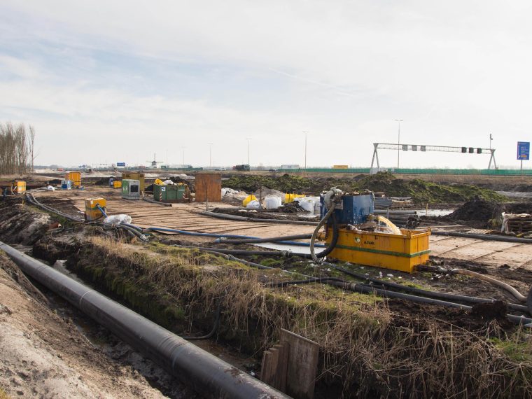 Bouwterrein met pompen, slangen en een leiding in een sloot, met een snelweg en molen op de achtergrond