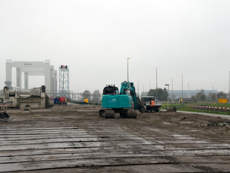 Graafmachine op rijplaten bij een bouwplaats, met de Botlekbrug op de achtergrond