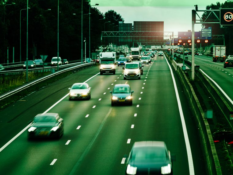 Druk verkeer met vrachtwagens en auto’s op een snelweg, onder matrixborden die 80 aangeven