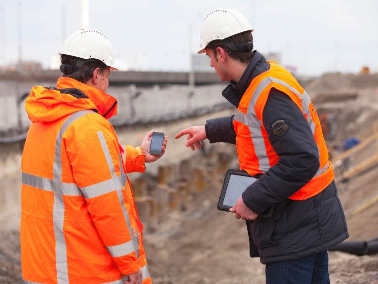 Twee medewerkers met helm en oranje veiligheidskleding bekijken een smartphone bij een bouwput
