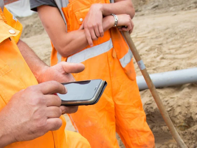 Grondwerker in oranje werkkleding gebruikt een smartphone naast een collega met een schop bij een sleuf met een buis
