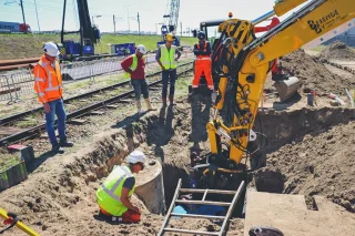 Grondwerkers in veiligheidskleding bij een open sleuf naast een spoorlijn, met een gele graafmachine