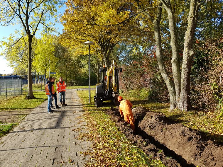Grondwerker in oranje overall graaft een sleuf voor kabels en leidingen langs een voetpad, terwijl 3 collega’s in veiligheidshesjes toekijken