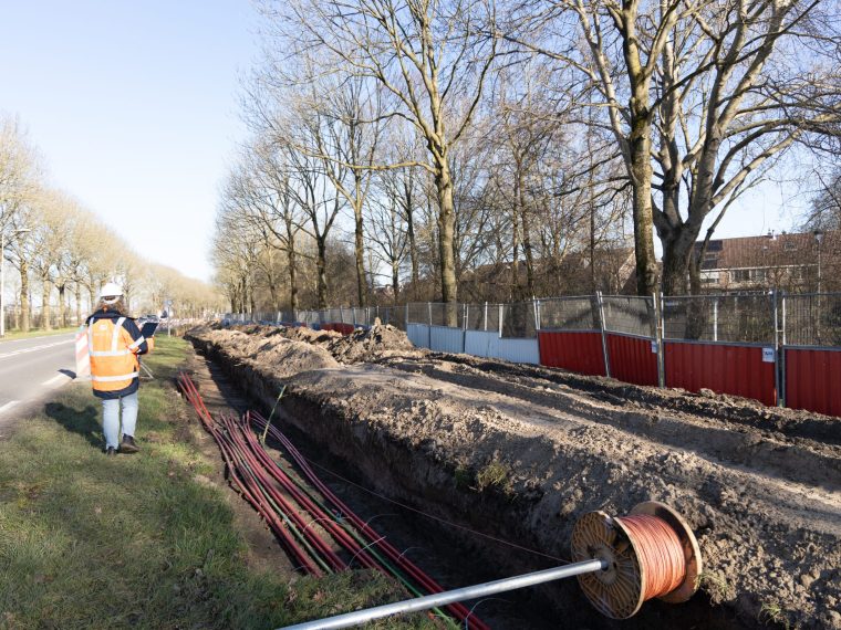 Medewerker in oranje veiligheidsjas met tablet bij een open sleuf met kabels en een kabelhaspel langs een weg